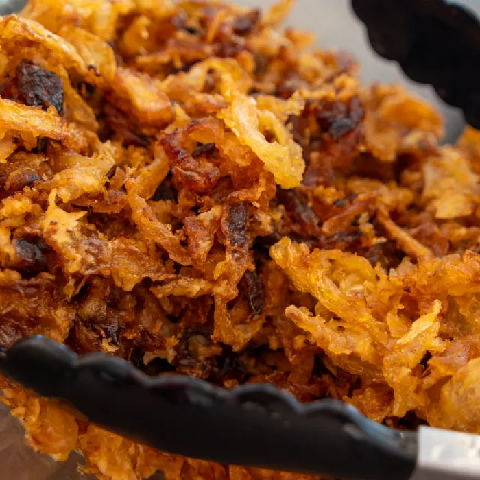 Frisch frittierte Röstzwiebeln in einer Glasschüssel, daneben eine Zange.