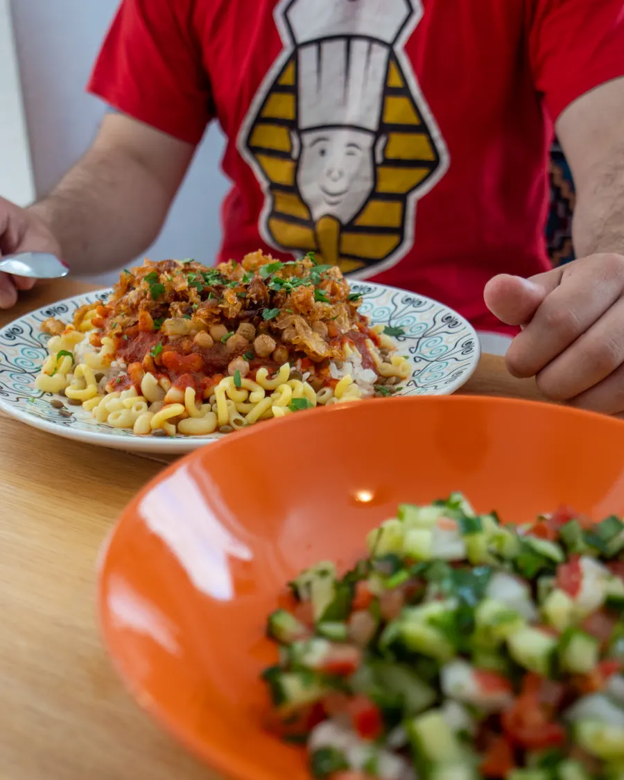 Am Tisch im Lokal, im roten Shirt mit dem Pharao-Logo, davor ein Teller Kuschari und der Kleopatra-Salat.