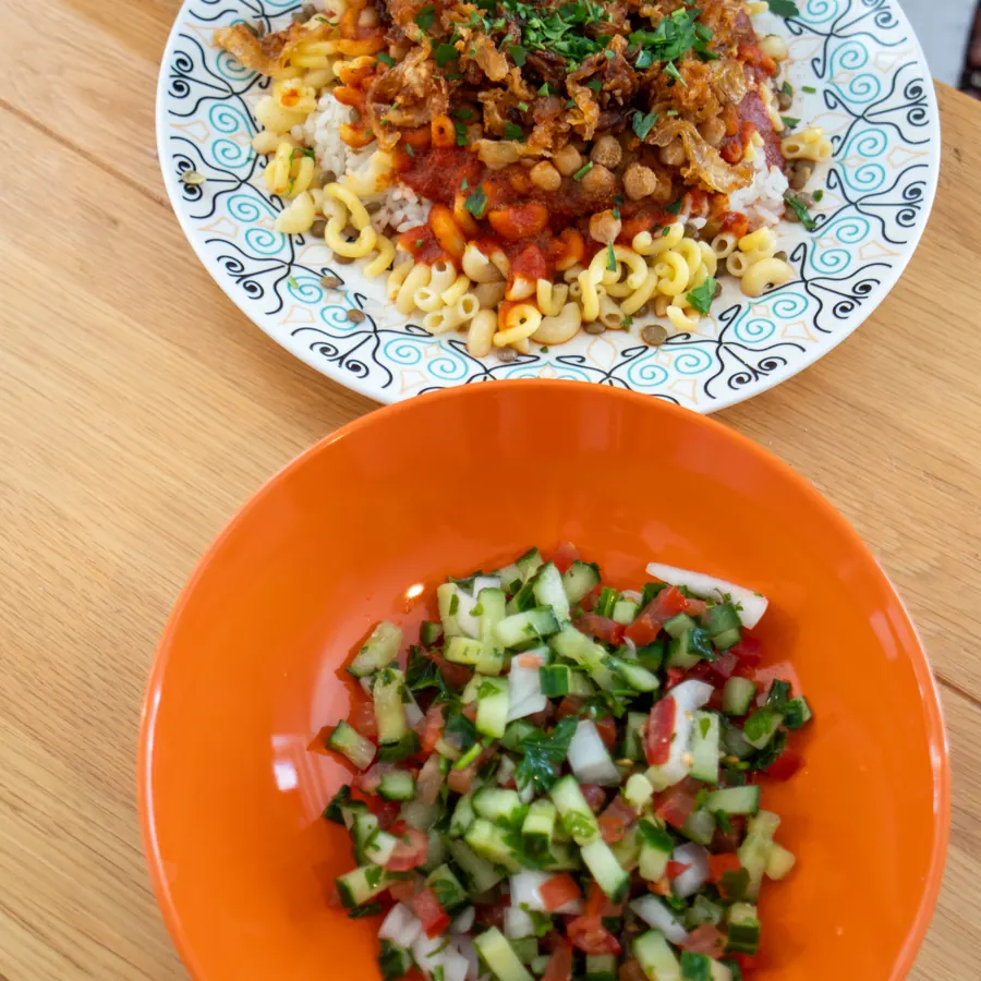 Ein Teller Kuschari und daneben eine orange Schale mit Kleopatra-Salat auf dem Holztisch.